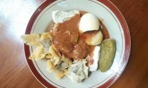 Sebuah piring batagor lezat yang dihidangkan di atas meja. Batagor adalah jajanan khas Bandung, terdiri dari bola ikan tenggiri yang dibalut dengan adonan tepung dan digoreng hingga renyah. Di gambar ini, batagor disajikan dengan tumpukan bakso ikan, irisan ketimun segar, dan ditaburi dengan bawang goreng dan kacang tanah. Saus kacang pedas dan kecap manis juga tersedia di sampingnya. Batagor adalah hidangan populer di Indonesia, terkenal karena rasa gurih dan tekstur renyahnya yang menggugah selera.